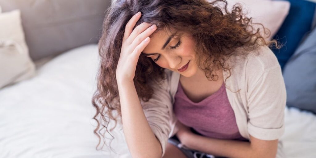 Young woman sitting on a bed, holding her stomach with a pained expression, conveying discomfort.